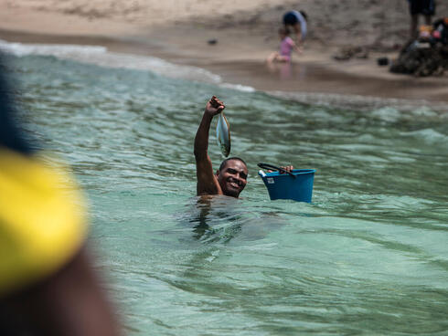 A man up to his neck in water smiles while holding up a fish and a blue bucket