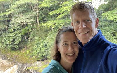A white man and woman take a selfie and smile into the camera in front of a lush green landscape.