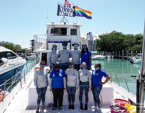 A group of people wearing face masks stand on the back of a boat and 