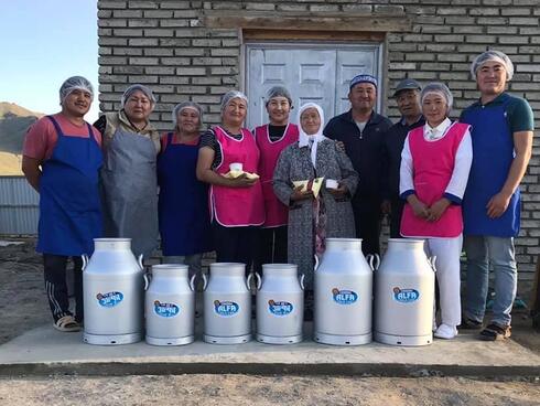 A group of people wearing blue or pink aprons and hair nets stand in a row smiling at the camera behind a row of metal milk jugs