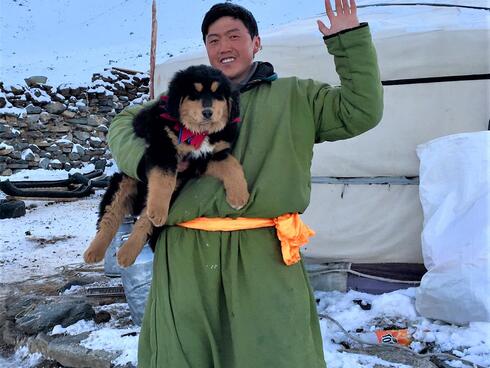 A man in a long green coat holds a black and white puppy and waves at the camera