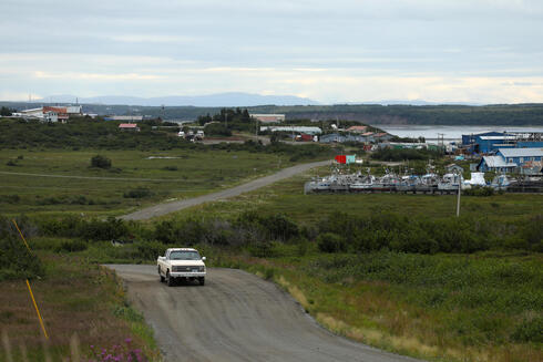 A car drives up a road with the village of Naknek in the background