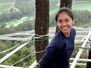 Nety Riana Sari stands beside a tree overlooking a forest and smiles at the camera