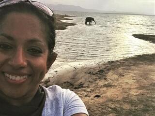 Nilanga Jayasinghe smiling at the camera in front of water with an elephant walking by