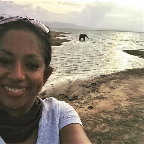 Nilanga Jayasinghe smiling at the camera in front of water with an elephant walking by