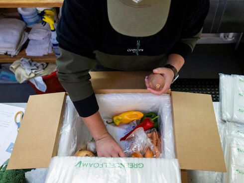 A person leans over a box of vegetables that they are packing to mail