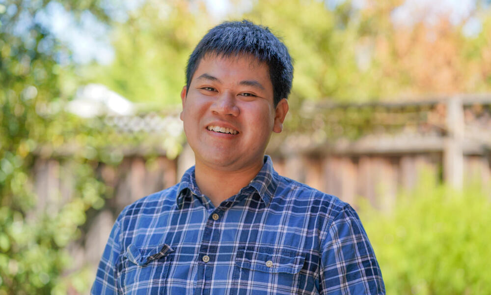 Headshot of Peter Pham in a blue plaid shirt smiling at the camera with greenery in the background