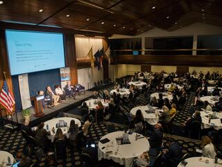 an aerial view of a large meeting room with people at tables watching a presentation on stage