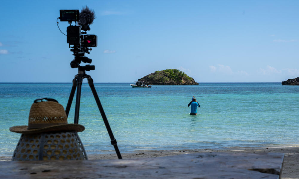 A snorkeler wades out into the calm, blue ocean at Raja Ampat. There is a camera on a tripod recording the scene in the foreground. 