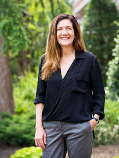 Rebecca shaw stands in a lush green setting. She has one hand in her pocket and is wearing a black top and grey slacks.