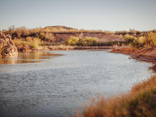 A calm stretch of river in dimming light with desert shoreline and blue sky