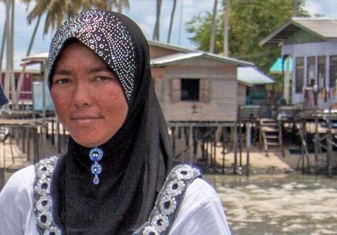 A portrait of a woman standing on a dock wearing a black headdress and white tshirt with a village on stilts in the background on the shoreline