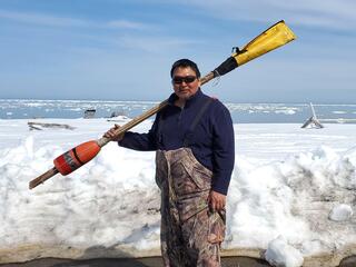A man wearing sunglasses holds a pole with an orange buoy at one end and a yellow cover at the other in front of the water