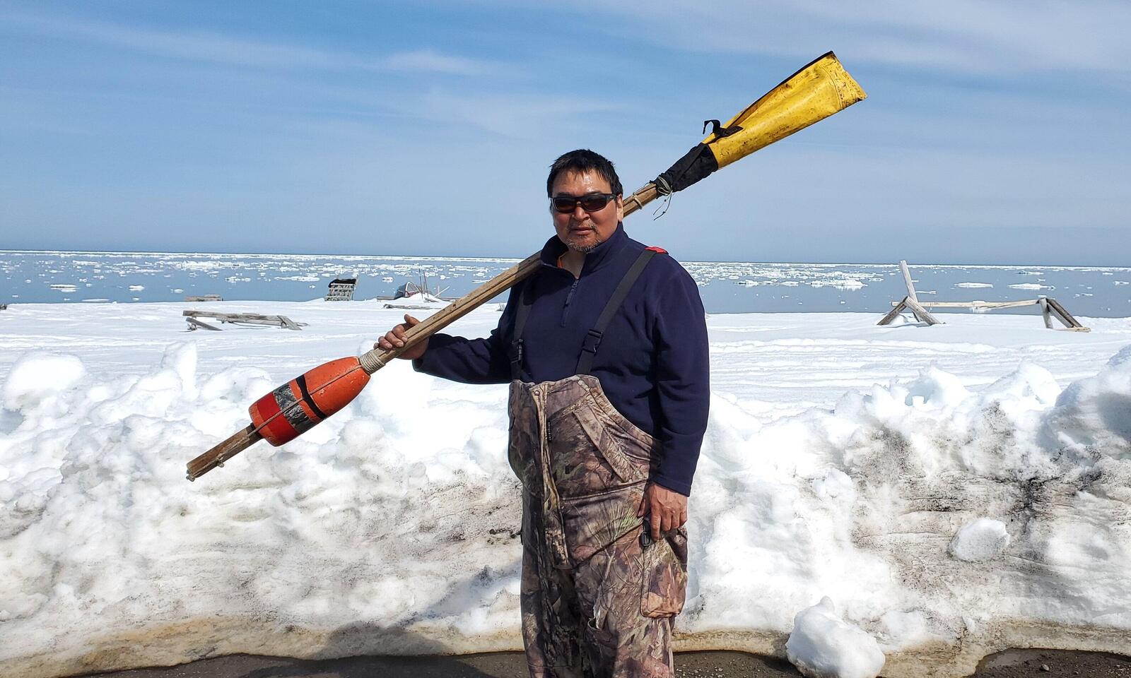 A man wearing sunglasses holds a pole with an orange buoy at one end and a yellow cover at the other in front of the water