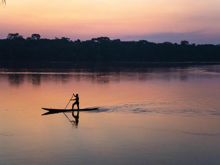 Someone standing up paddling on a slim canoe along a river at sunset with pink and purple hues