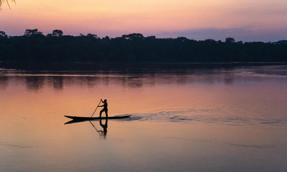 Someone standing up paddling on a slim canoe along a river at sunset with pink and purple hues