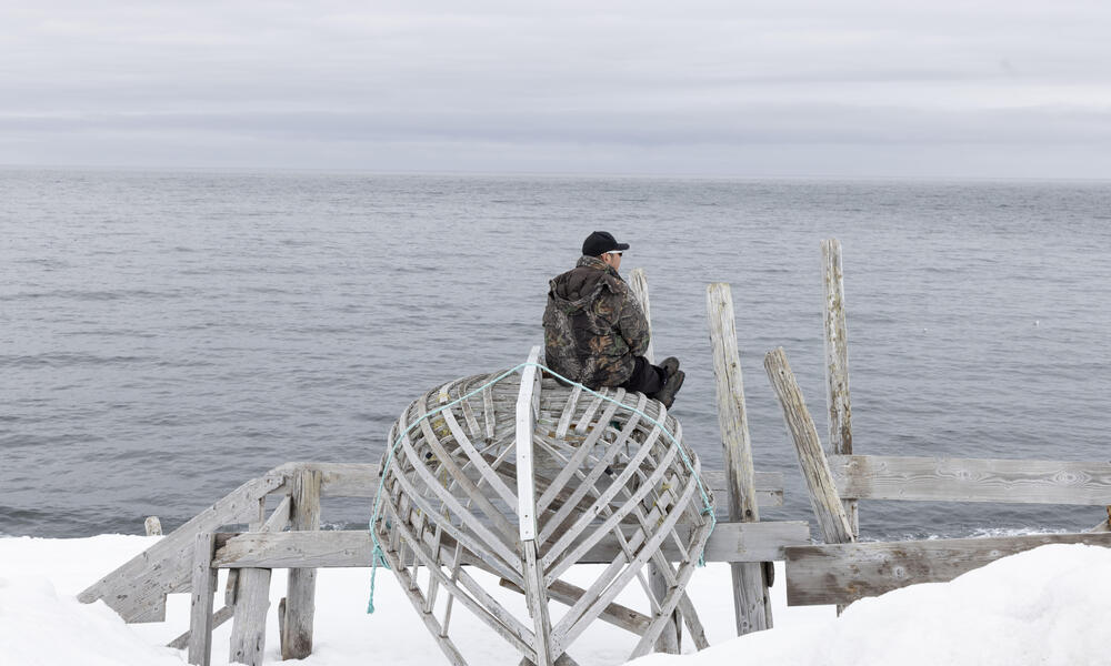 A person wearing a black jacket sits on top of an upsidedown boat looking out at the sea