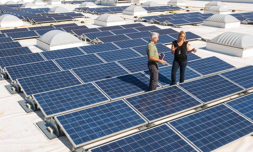 Two people stand on a roof that is covered with solar panels
