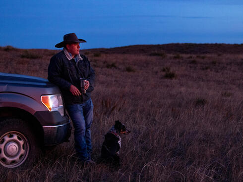 TJ Heinert stands with his dog Bandita at the Wolakota Buffalo Range on the Rosebud Sioux Reservation in South Dakota under a dimly lit sky