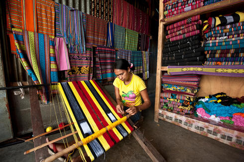 A woman stands at a loom with colorful fabrics on shelves behind her