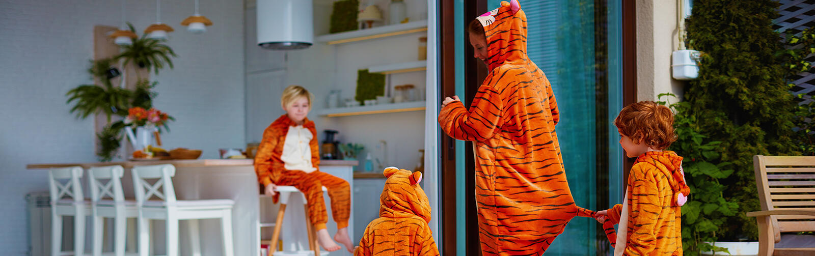 A young family with three children wear tiger onesies. The adult is walking into the apartment, while two children follow. The third child sits on a high top chair smiling at them.