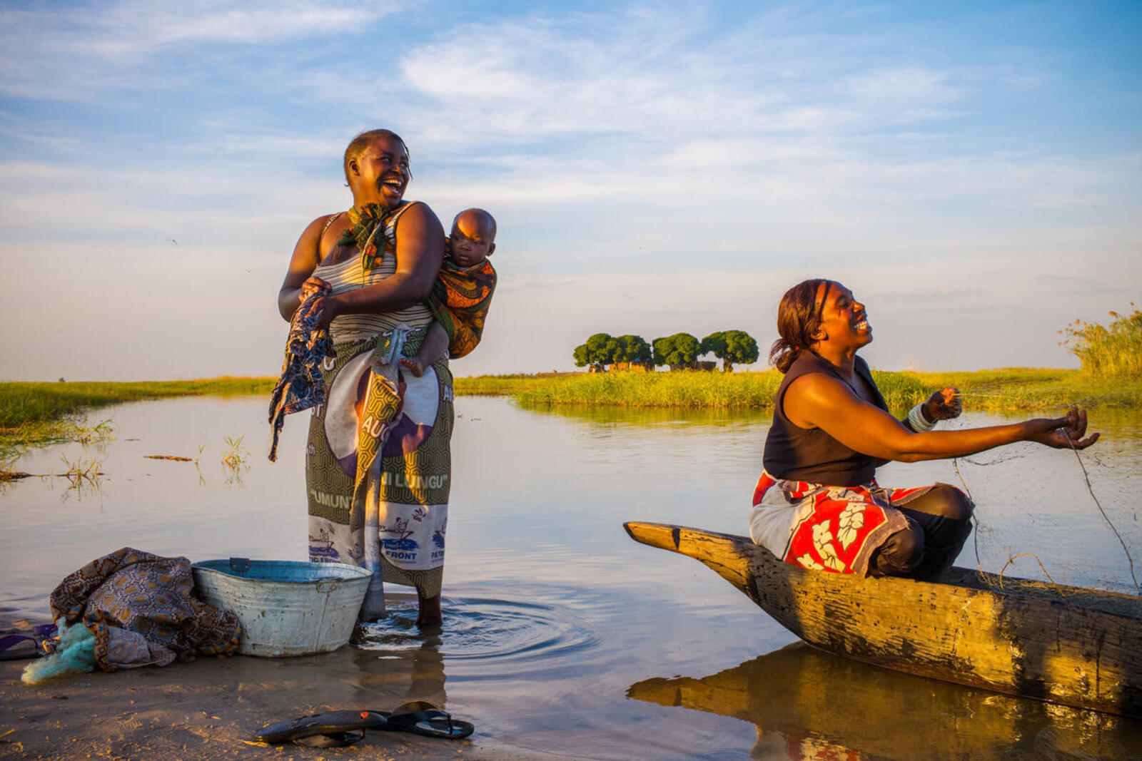 Two women and a baby on a river bank, one washing clothes and the other casting a net while all smile