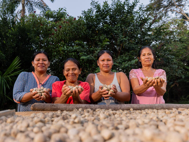 Four women stand in front of a tray full of seeds and each hold a two handfuls of seeds and smile at the camera.
