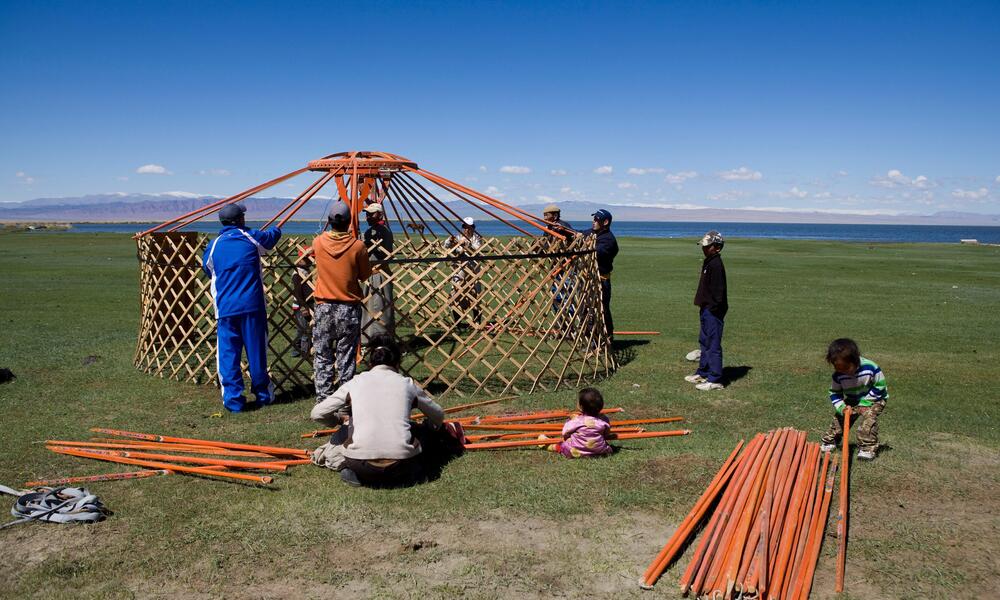 A group of people build a yurt with lattice wood walls on a grassy landscape