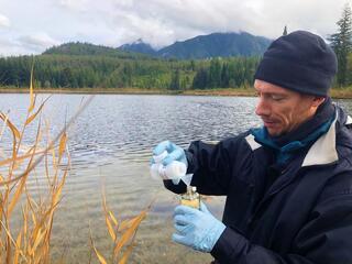 WWF scientist is shown with water sampling device in front of water and mountain backdrop in British Columbia