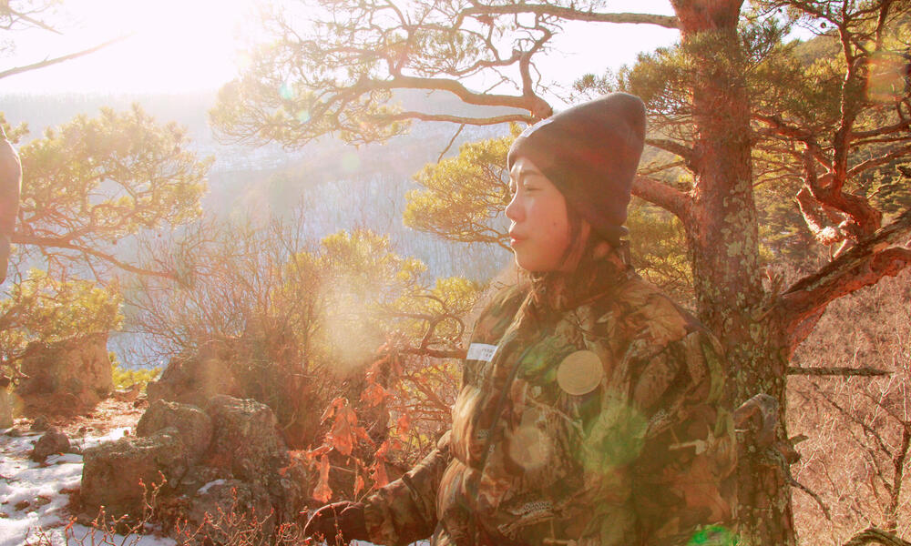 A female ranger prepares to begin her day, standing against a forest backdrop as the sun rises