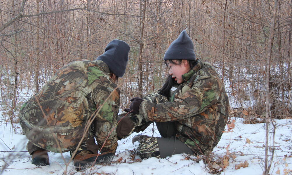Two female rangers sit in the forest while one helps the other remove her foot from a snare
