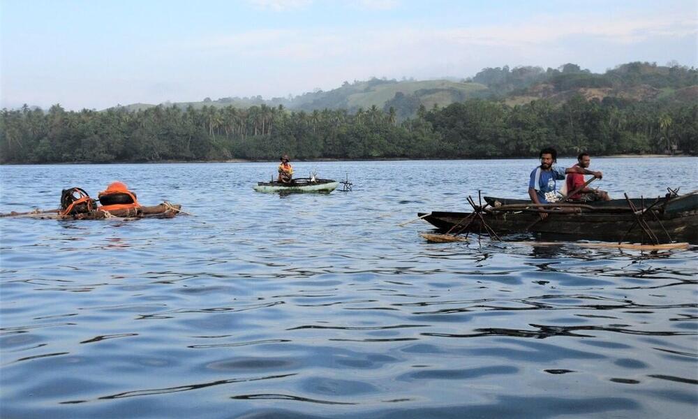 men on small wooden boats paddle across calm blue water with a coastline of trees in the background