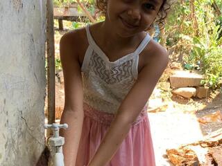 A girl smiles into the camera as she washes her hands under an outdoor faucet