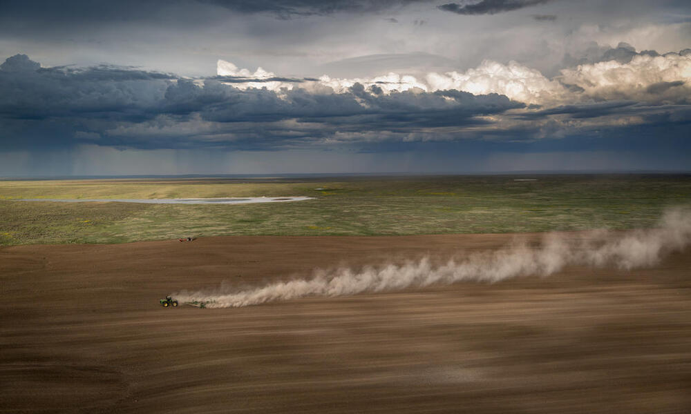 A tractor plows a vast field with a rain storm in the distant background
