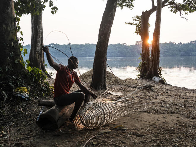 A man sitting on a riverbank weaving a fishing net out of reeds at sunset