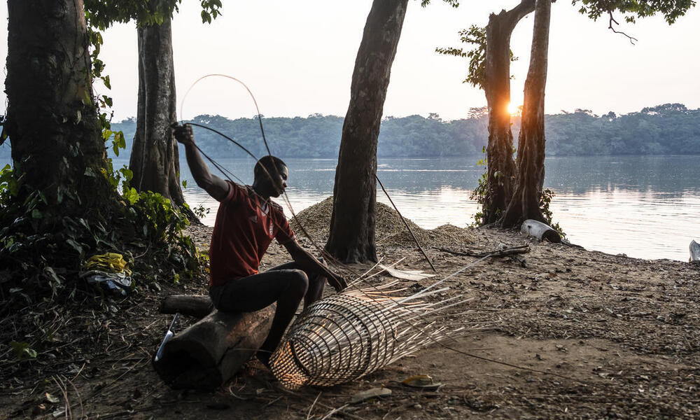 A man sitting on a riverbank weaving a fishing net out of reeds at sunset