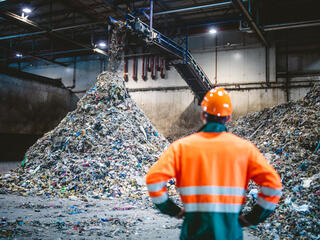 Rear view of young male worker in helmet, pollution mask, and reflective clothing observing waste falling from conveyor belt onto pile at facility.