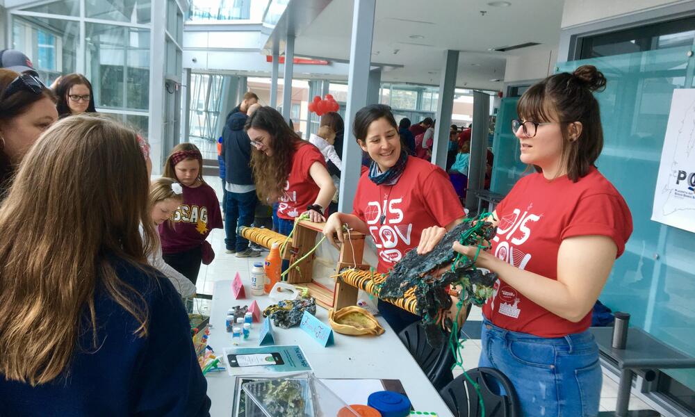 three women in red tshirts standing behind white folding tables showing passersby their educational materials