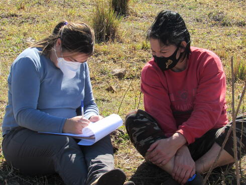Two women sit on the grassy ground, one holds a notepad taking notes and each wear face masks