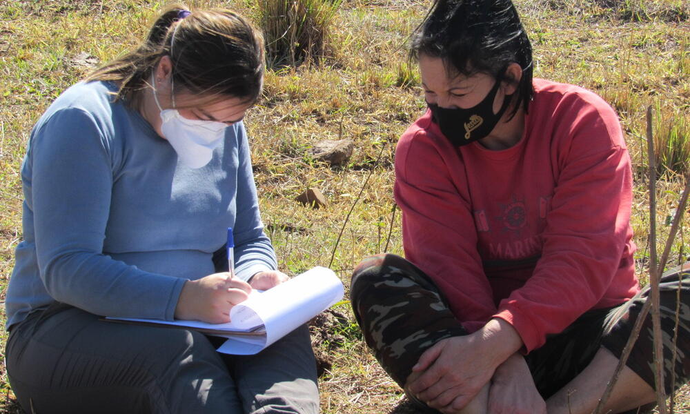 Two women sit on the grassy ground, one holds a notepad taking notes and each wear face masks