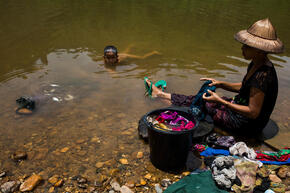 A woman sits on a river bank washing clothes while a child swims naked in the water