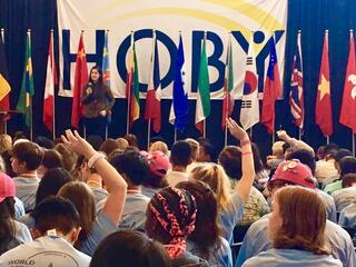 A woman standing on a stage in front of a row of flags, presenting to an audience of children with their hands up