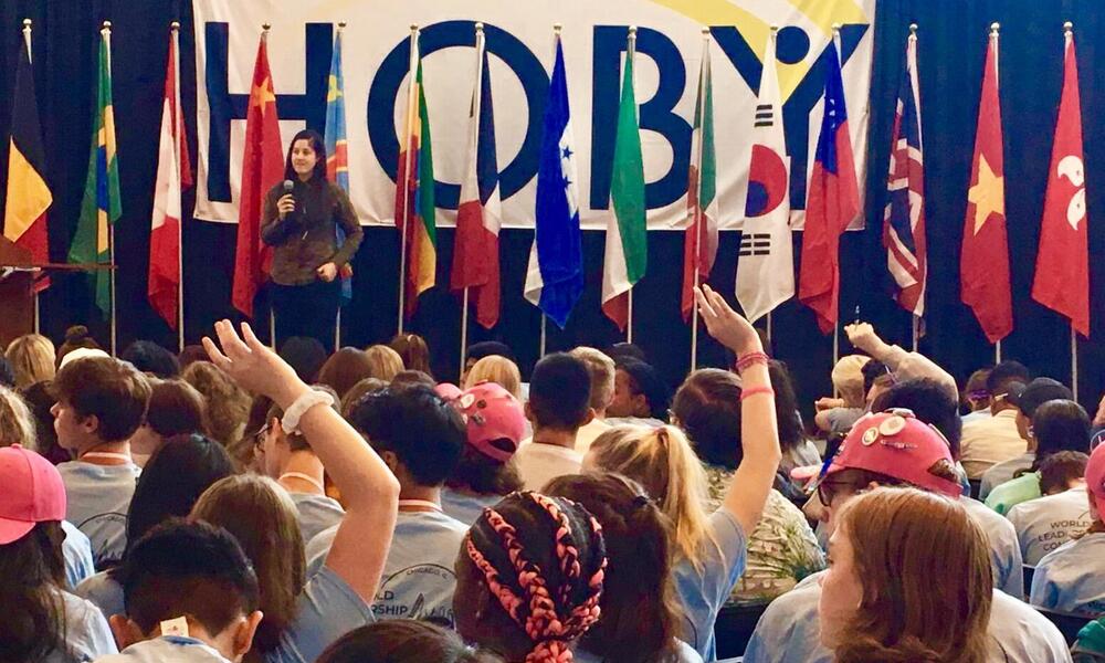 A woman standing on a stage in front of a row of flags, presenting to an audience of children with their hands up